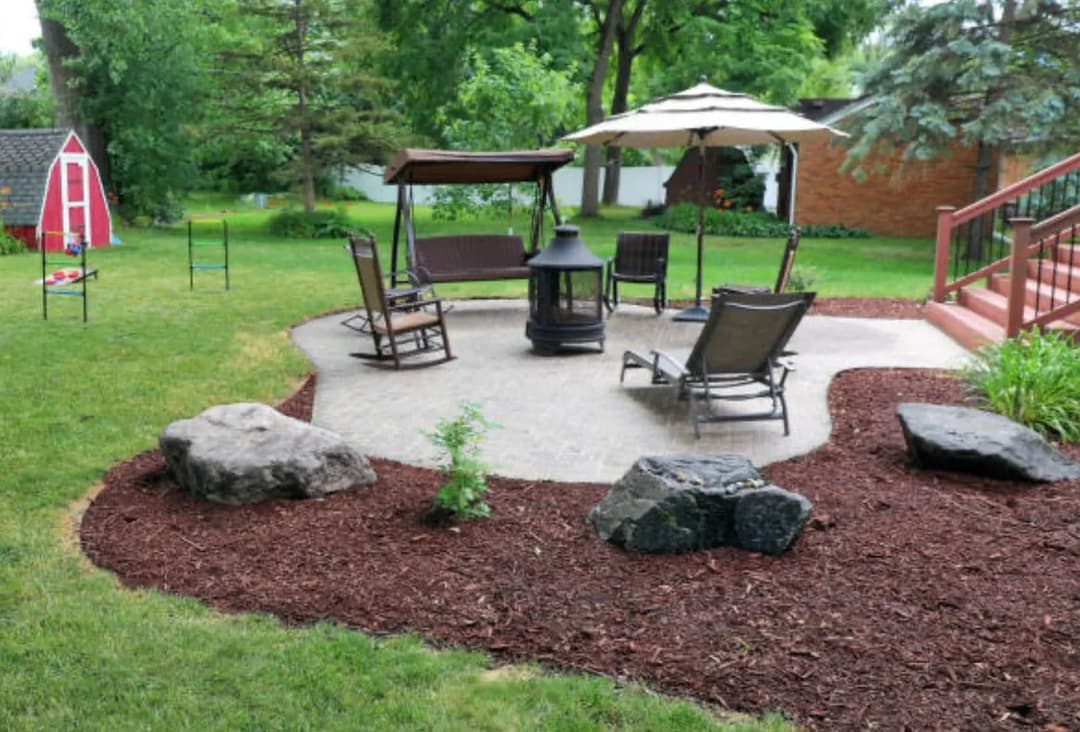 Patio seating area with rocking chairs, a fire pit, and landscaping in a green backyard.
