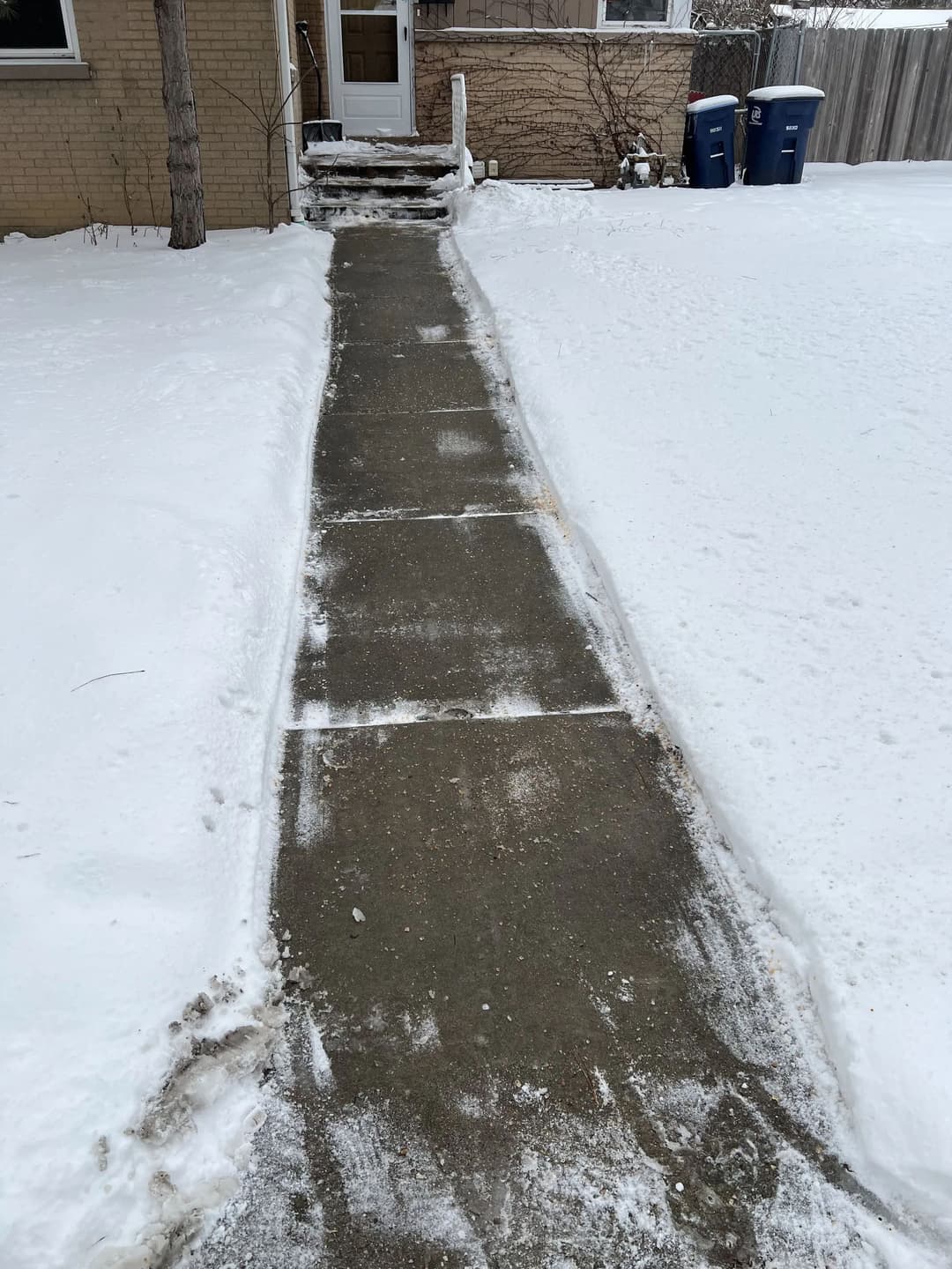 Snow-covered sidewalk leading to a house entrance with cleared path and trash bins nearby.