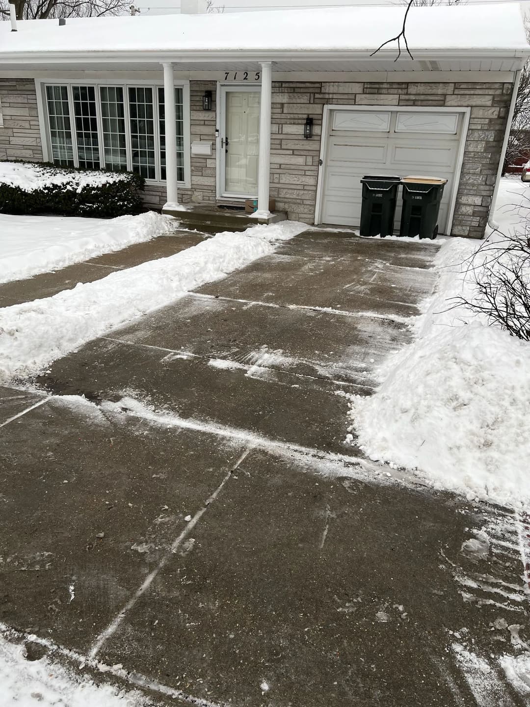 Cleared driveway at house 7123 with snow piles and trash bins on a winter day.