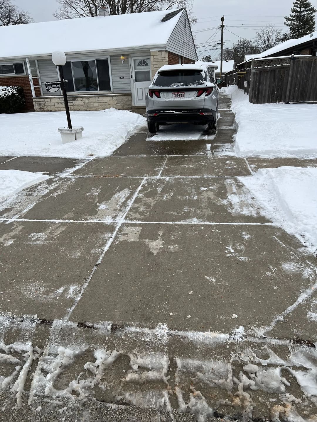Snow-covered driveway with cleared path, residential home, and parked vehicle in winter scene.