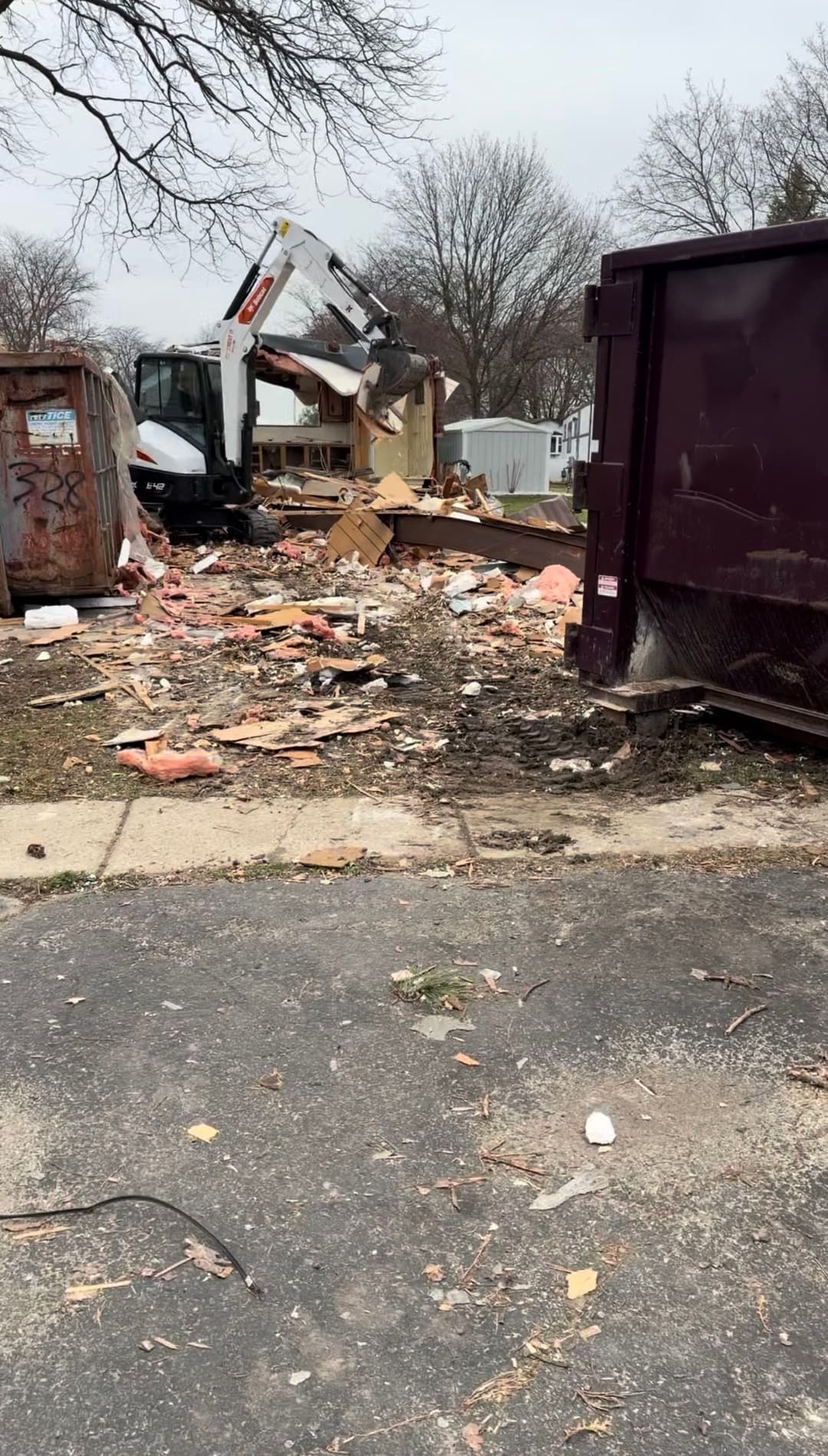 Excavator demolishing a building amid debris and a dumpster on a cloudy day.