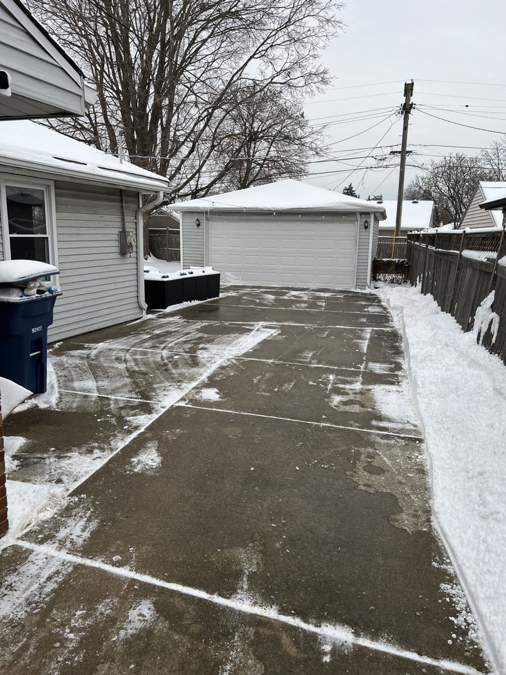 Snow-cleared driveway with nearby garage and blue trash bin in winter setting.