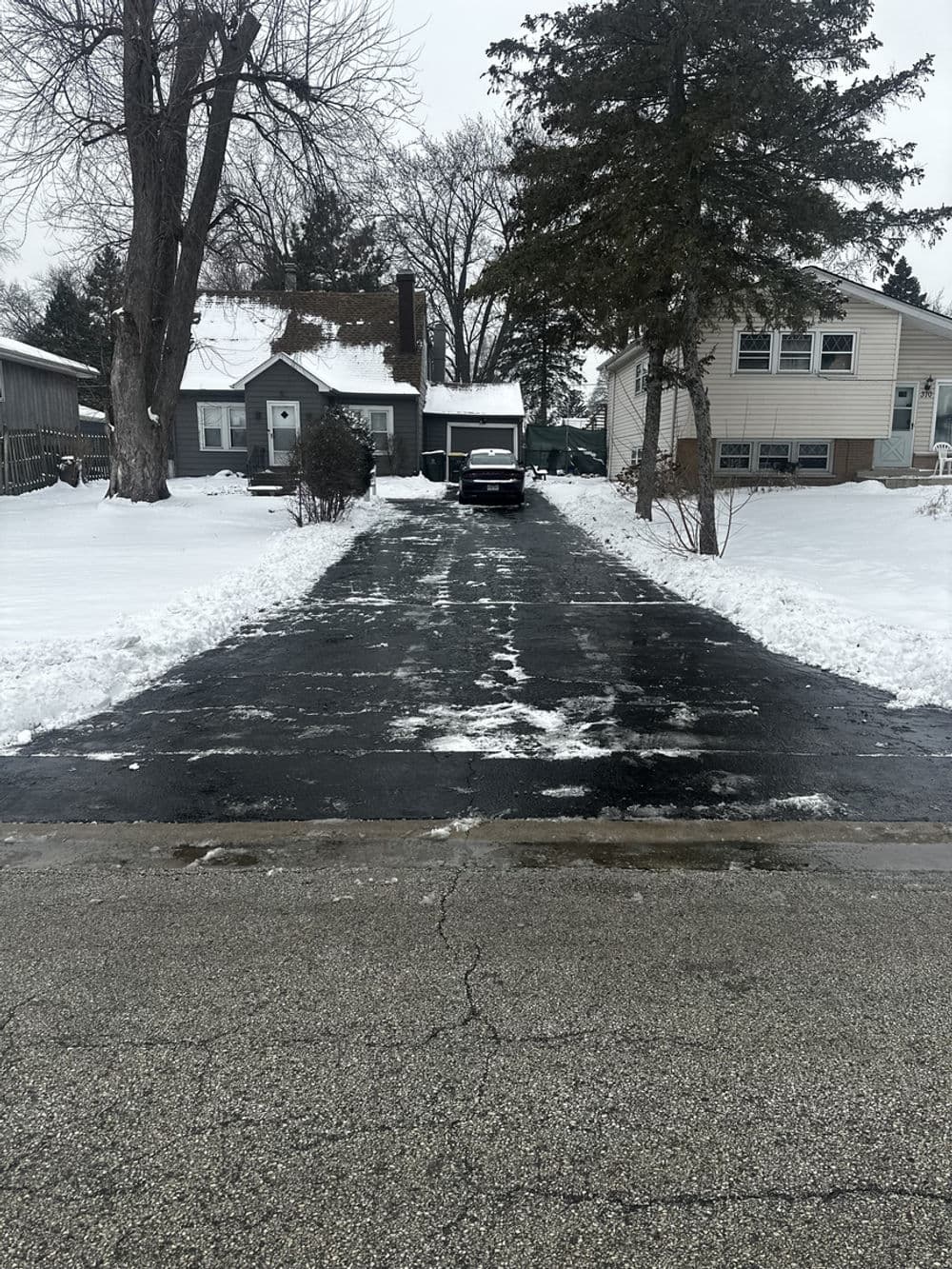 Snow-covered driveway in a residential area with two houses and winter trees.