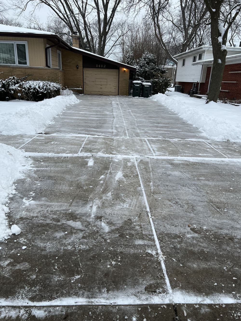 Cleared snow driveway with marked sections and residential homes in background on winter day.