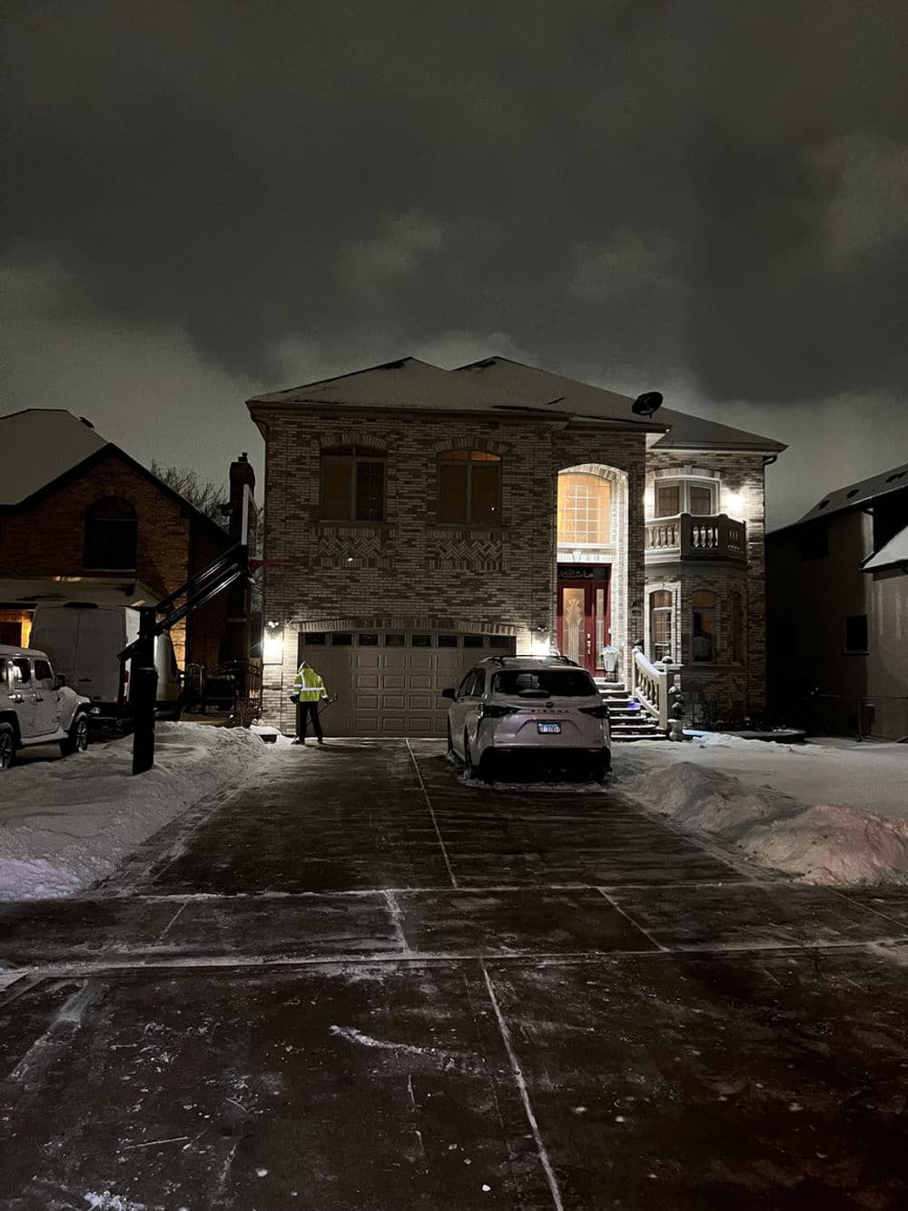 Snowy night scene of a two-story brick house with a car in the driveway and a person standing outside.