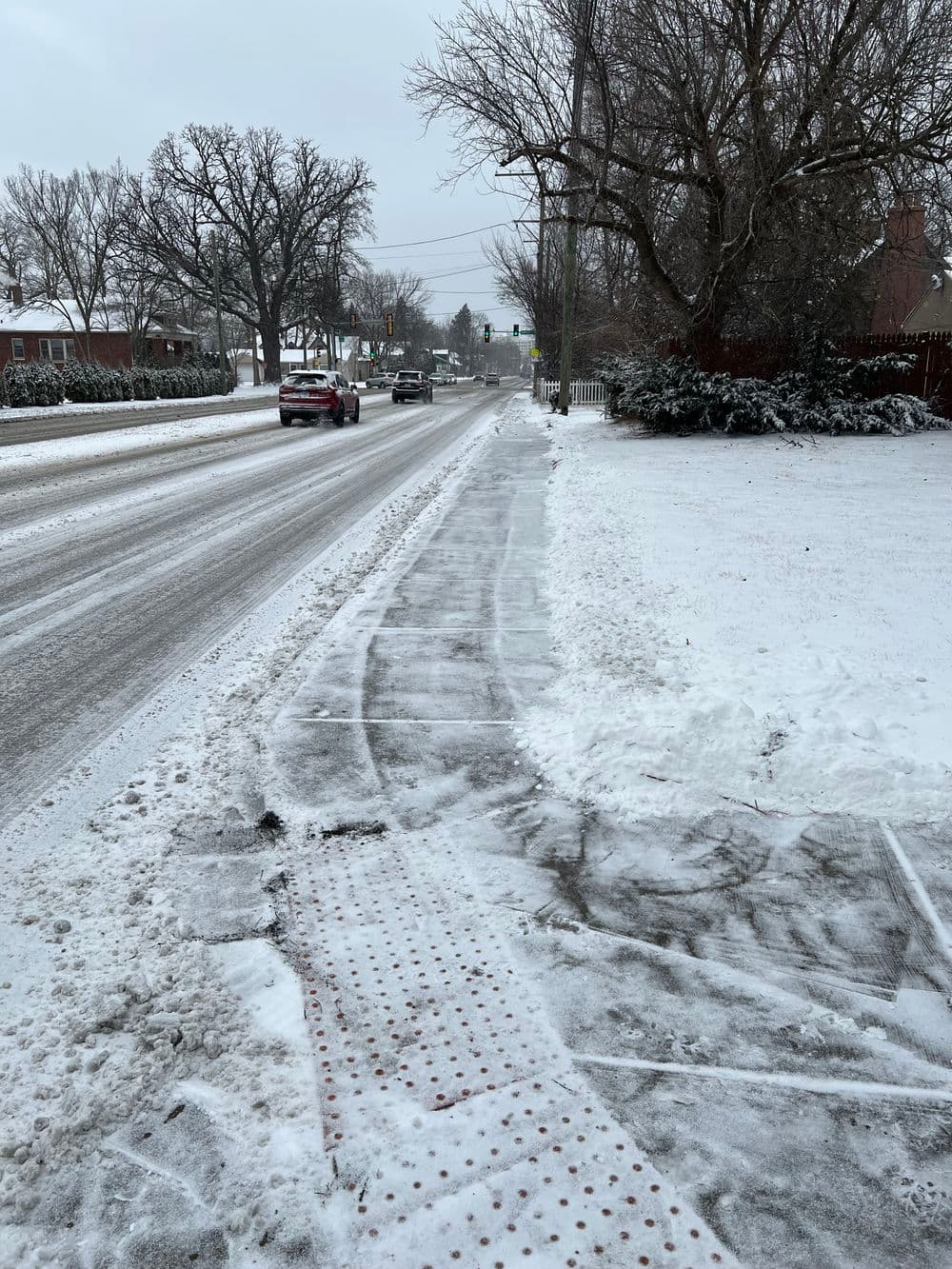 Snowy sidewalk along a road with sparse traffic and winter trees in a quiet neighborhood.