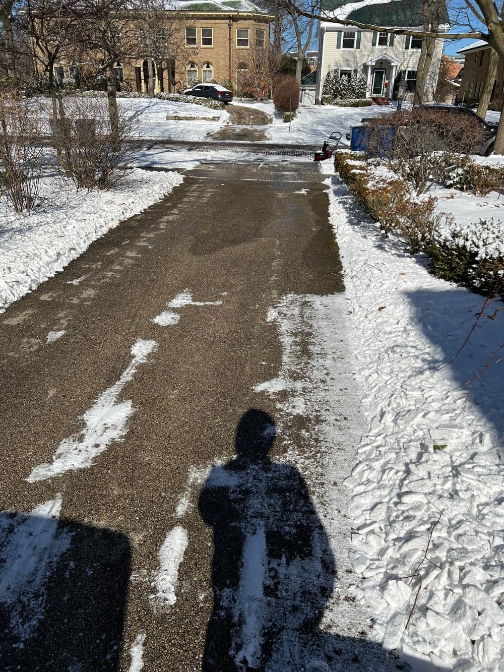 Snow-covered driveway with clear path, residential houses in the background, and winter scenery.